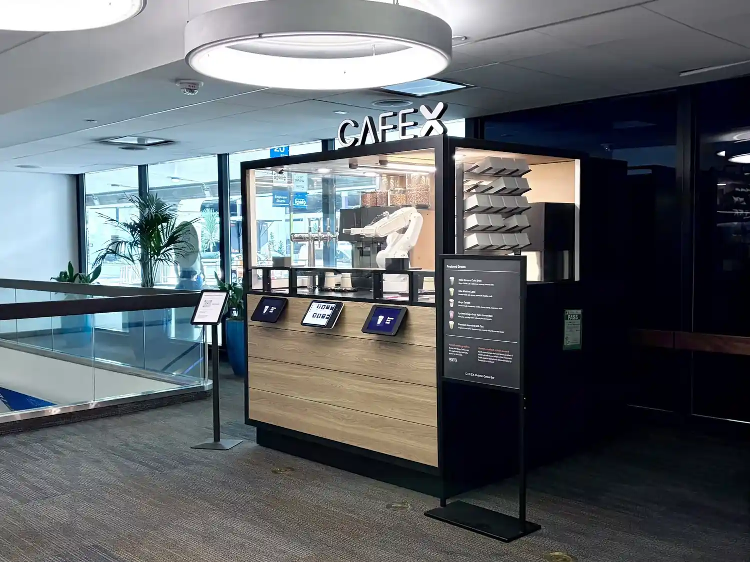The Cafe X robotic coffee kiosk at SFO — a glass-walled booth with a white robot arm, order tablets on the front, and the CAFE X sign lit overhead