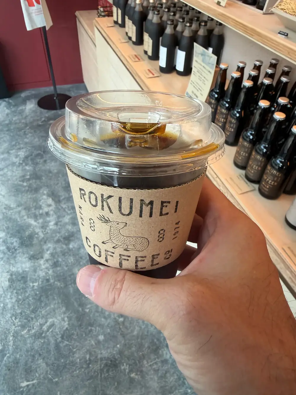 An iced Rokumei Coffee cup in hand at the Shibuya Scramble Square bar, coffee bottles lining the counter behind it